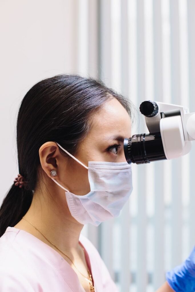 Dentist in protective mask using a microscope for precise dental examination.