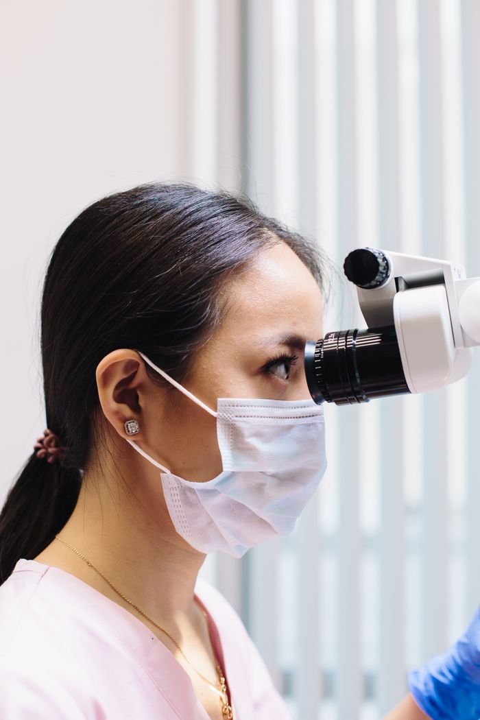 Dentist in protective mask using a microscope for precise dental examination.