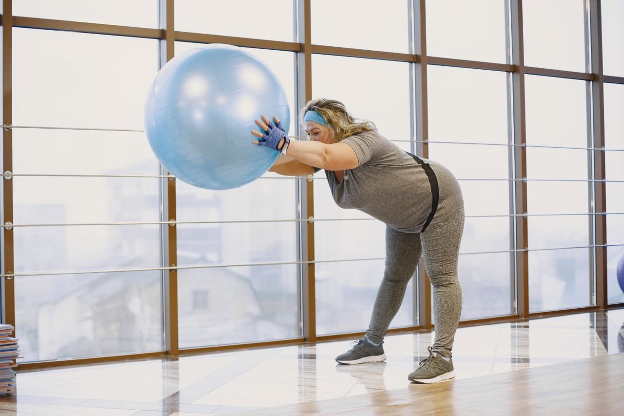 ÜROJİNEKOLOJİ A plus size woman stretches using a fitness ball in a bright gym, promoting wellbeing and body positivity.
