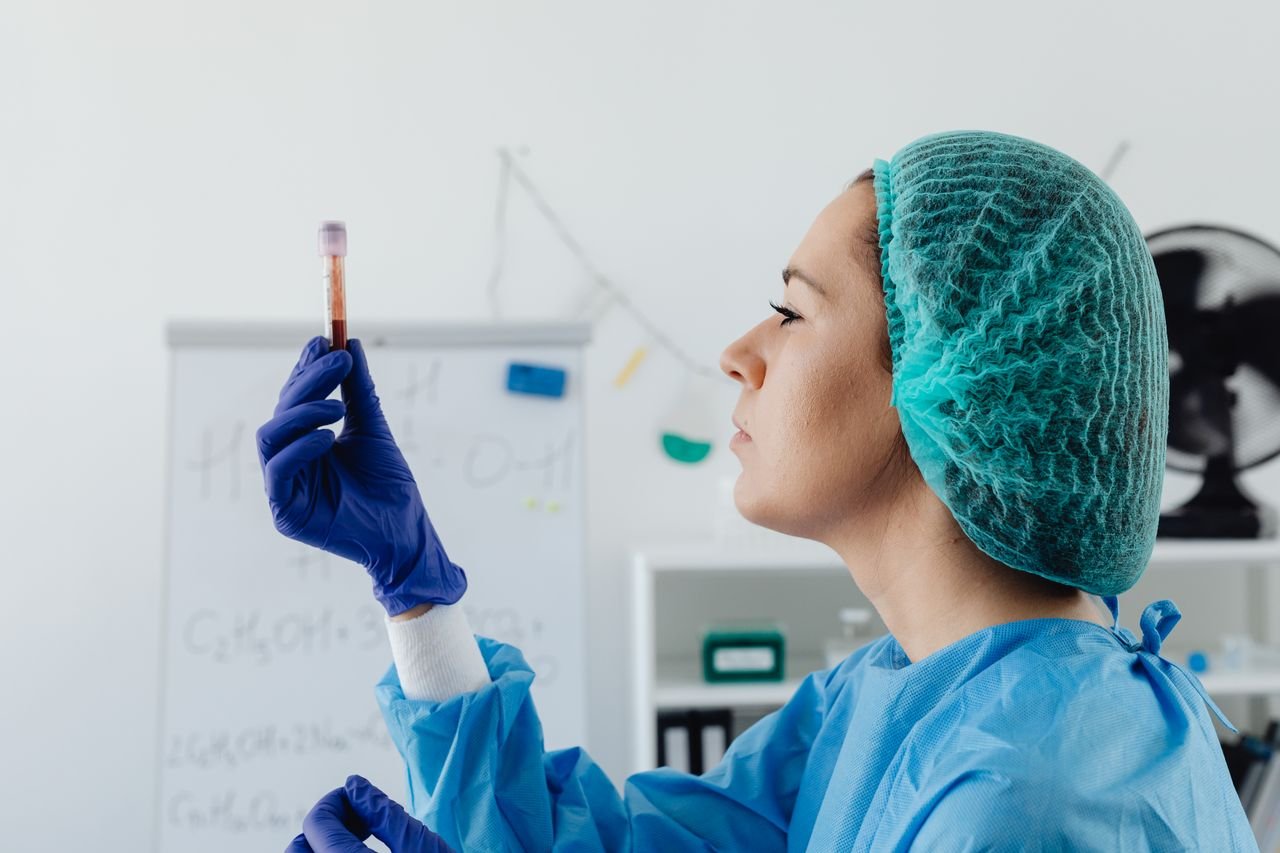 A female scientist in protective gear inspects a blood sample vial in a lab setting.