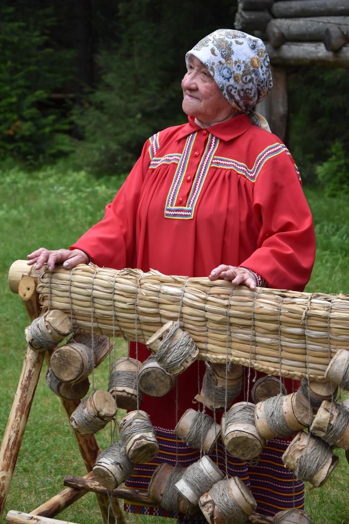 ÜROJİNEKOLOJİ Senior woman in traditional Russian attire stands outdoors with birch bark crafts. Rural cultural scene.